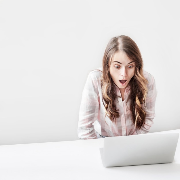Mujer joven confundida sentada en la mesa y usando una computadora portátil en un fondo gris