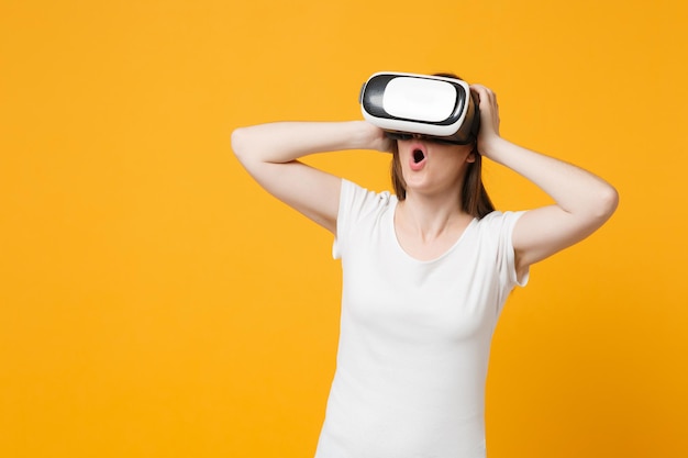 Mujer joven conmocionada con ropa blanca informal mirando con auriculares de realidad virtual, poniendo las manos en la cabeza aislada en un fondo naranja amarillo brillante. Concepto de estilo de vida de las personas. Simulacros de espacio de copia.