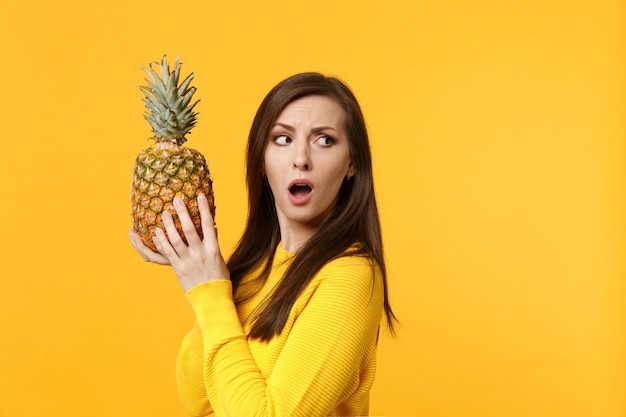 Mujer joven conmocionada con ropa informal mirando a un lado, sostenga fruta fresca de piña madura aislada en un fondo amarillo anaranjado en el estudio. Estilo de vida vivo de la gente, concepto de vacaciones relajantes. Simulacros de espacio de copia