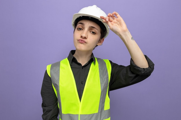 Mujer joven constructor en chaleco de construcción y casco de seguridad mirando con expresión seria y segura tocando su casco