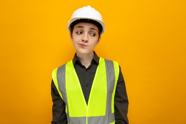 Mujer joven constructor en chaleco de construcción y casco de seguridad mirando a un lado con expresión escéptica de pie sobre la pared naranja