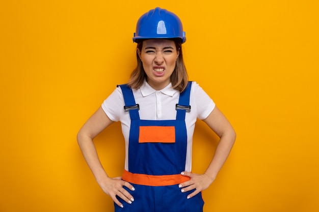 Mujer joven constructor en uniforme de construcción y casco de seguridad con cara enojada con los brazos en la cadera de pie en naranja