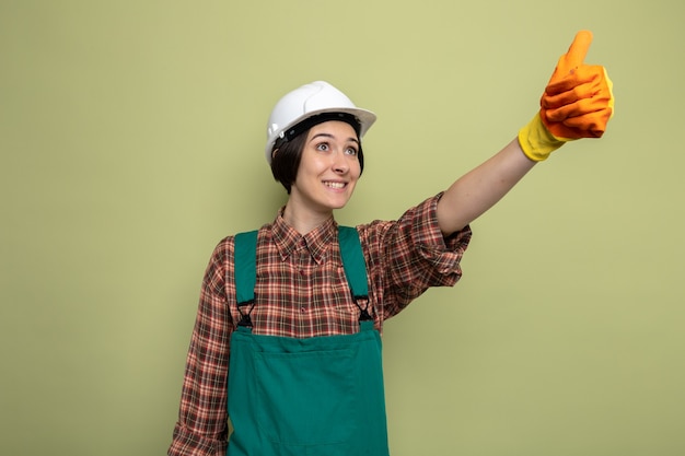 Mujer joven constructor en uniforme de construcción y casco de seguridad en guantes de goma mirando hacia arriba sonriendo alegremente mostrando los pulgares para arriba de pie en verde