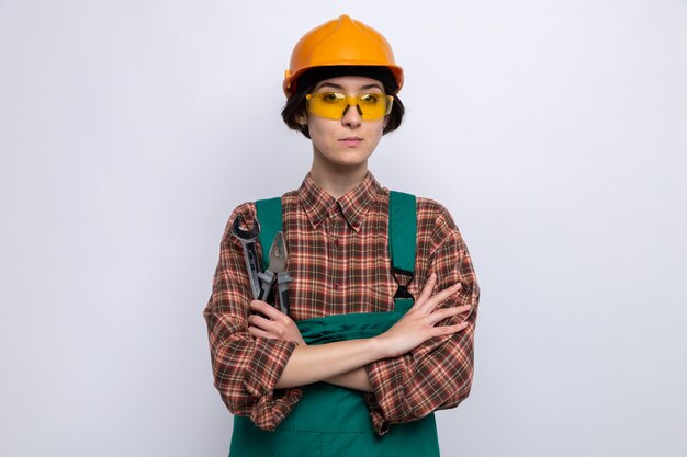 Mujer joven constructor en uniforme de construcción y casco de seguridad con llave con alicates con expresión seria de confianza con los brazos cruzados de pie sobre la pared blanca