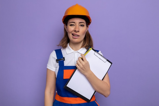 Mujer joven constructor en uniforme de construcción y casco de seguridad sosteniendo portapapeles con páginas en blanco con sonrisa escéptica en la cara de pie sobre la pared púrpura