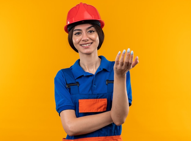 Mujer joven constructora complacida en uniforme extendiendo la mano aislada en la pared amarilla