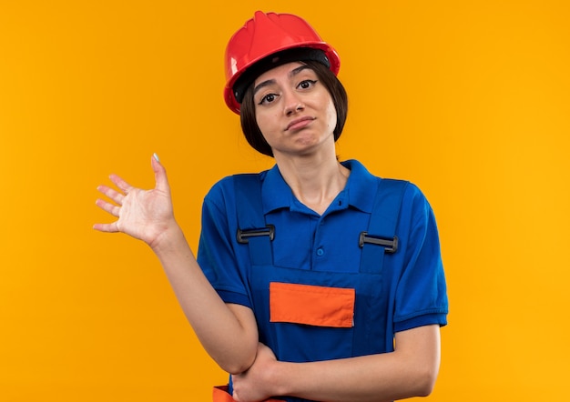 Mujer joven constructora disgustada en uniforme extendiendo la mano aislada en la pared amarilla