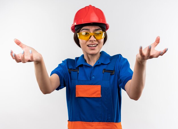 Mujer joven constructora disgustada en uniforme con gafas tendiendo las manos a la cámara