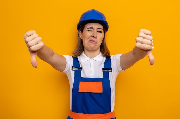 Mujer joven constructora insatisfecha en uniforme de construcción y casco de seguridad mostrando los pulgares hacia abajo de pie sobre la pared naranja