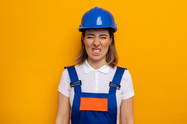 Mujer joven constructora en uniforme de construcción y casco de seguridad mirando molesto e irritado haciendo boca torcida de pie en naranja