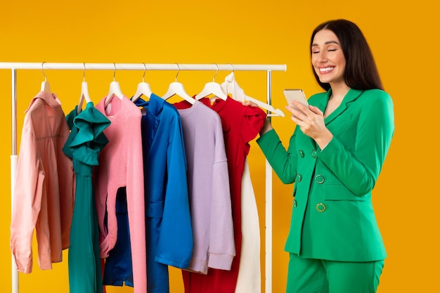 Mujer joven contenta adicta a las compras con compras de teléfonos celulares mientras está de pie cerca del riel de ropa eligiendo ropa de moda