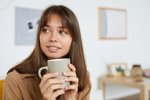 Mujer joven contenta sonriente con cabello castaño mirando hacia otro lado y bebiendo té de la taza en casa