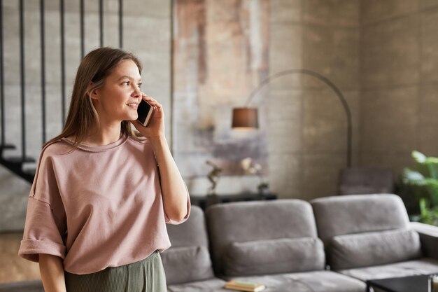 Foto mujer joven conversando por teléfono móvil y sonriendo mientras está de pie en la habitación en casa