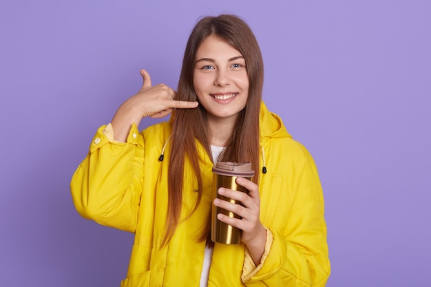 Mujer joven coqueteando haciendo gesto de teléfono como dice que me llamen aislado sobre fondo lila, sosteniendo una taza termo con bebida caliente, mira a la cámara sonriendo, vistiendo una chaqueta amarilla.