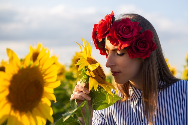 Mujer joven en una corona de flores rojas huele un girasol en un campo de girasoles al atardecer.