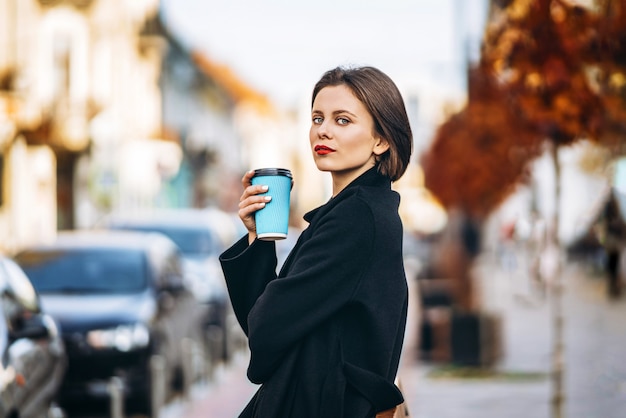 Mujer joven con corte de pelo corto y labios rojos, sostiene una taza de café, paseando por las calles de la ciudad. Alrededor hay personas y autos.