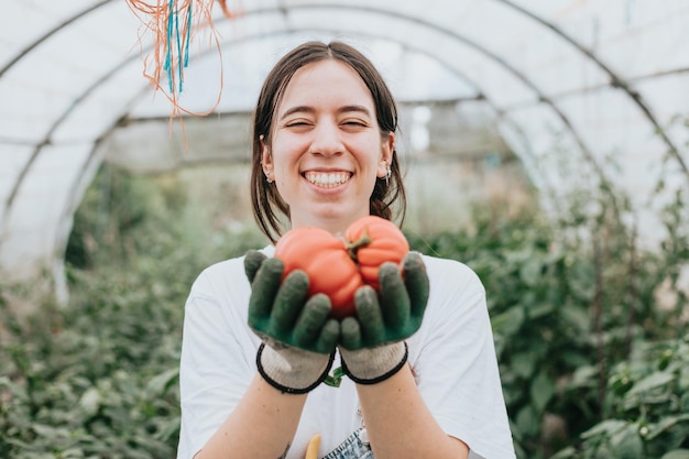 Mujer joven cosechando tomates frescos del invernadero mientras usa guantes y muestra a la cámara Sostenibilidad y concepto de alimentos saludables Productos crudos orgánicos cultivados en una granja doméstica