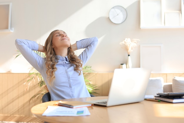 Mujer joven cruzó las manos detrás de la cabeza, disfrutando del descanso en casa. Mujer de negocios tranquila y despreocupada descansando en la mesa con la computadora, mirando a un lado, soñando con el futuro