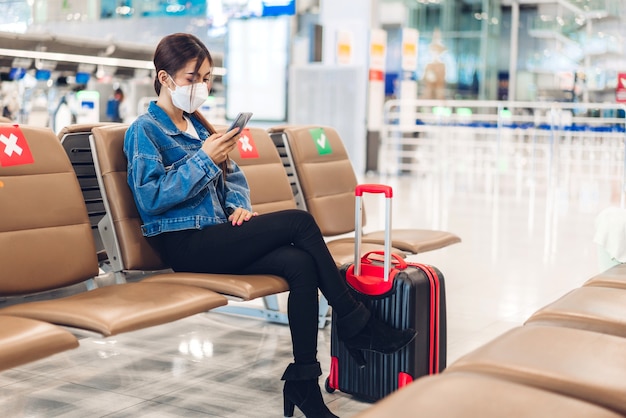 Mujer joven en cuarentena por coronavirus con máscara quirúrgica, protección facial durante la pandemia de virus con distanciamiento social, uso de teléfono inteligente escribiendo mensaje con equipaje, espera, vuelo, viaje en el aeropuerto