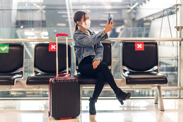 Mujer joven en cuarentena por coronavirus con máscara quirúrgica, protección facial durante la pandemia de virus con distanciamiento social, uso de teléfono inteligente escribiendo mensaje con equipaje, espera, vuelo, viaje en el aeropuerto