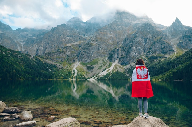 Mujer joven cubierta con la bandera de polonia mirando el lago en las montañas tatra