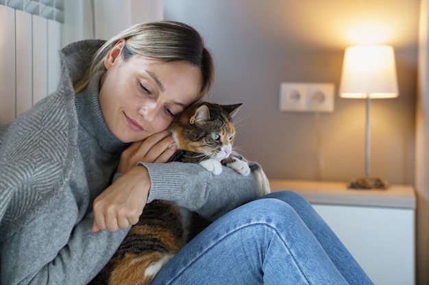 Mujer joven cubierta con una manta cálida se siente mejor abrazando a un gato doméstico e intenta continuar con la vida