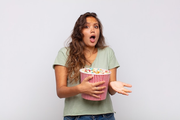 Mujer joven con un cubo de pop conrs que se siente extremadamente conmocionada y sorprendida, ansiosa y en pánico, con una mirada estresada y horrorizada