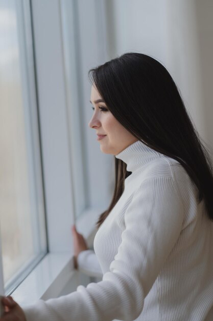 Mujer joven en cuello alto blanco mira la ventana pensando o soñando