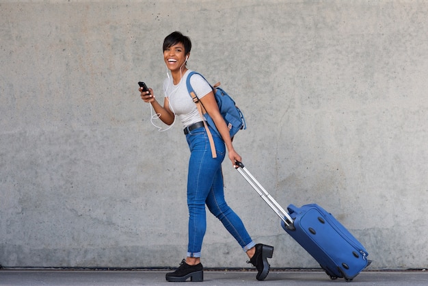 Mujer joven de cuerpo completo caminando con bolsa de viaje y teléfono móvil