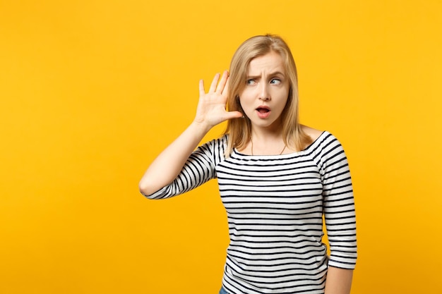 Mujer joven curiosa con ropa a rayas tratando de oírte, sosteniendo la mano cerca de la oreja aislada en el fondo de la pared naranja amarilla en el estudio. Emociones sinceras de la gente, concepto de estilo de vida. Simulacros de espacio de copia.