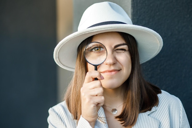 Mujer joven curiosa con sombrero blanco mirando a través de una lupa