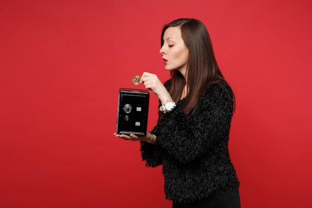 Mujer joven curiosa en un suéter de piel sosteniendo un banco de metal seguro para la acumulación de dinero, bitcoin, moneda futura aislada sobre fondo rojo. Personas sinceras emociones, concepto de estilo de vida. Simulacros de espacio de copia.