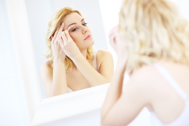 Mujer joven dar forma a las cejas con pinzas en el baño.