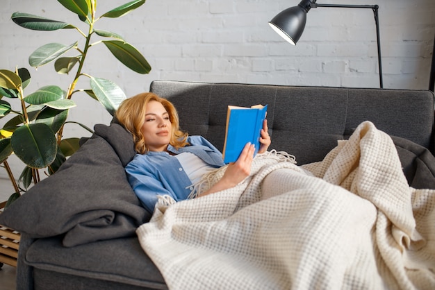 Mujer joven, debajo, un, manta, libro de lectura, en, sofá