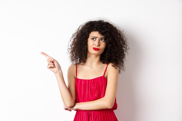 Mujer joven decepcionada y escéptica con el pelo rizado, vestido rojo, haciendo muecas y señalando con el dedo a la izquierda en el logo, de pie sobre fondo blanco.