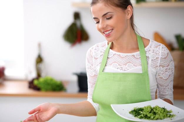 Mujer joven en delantal verde está cocinando en una cocina Ama de casa está ofreciendo ensalada fresca