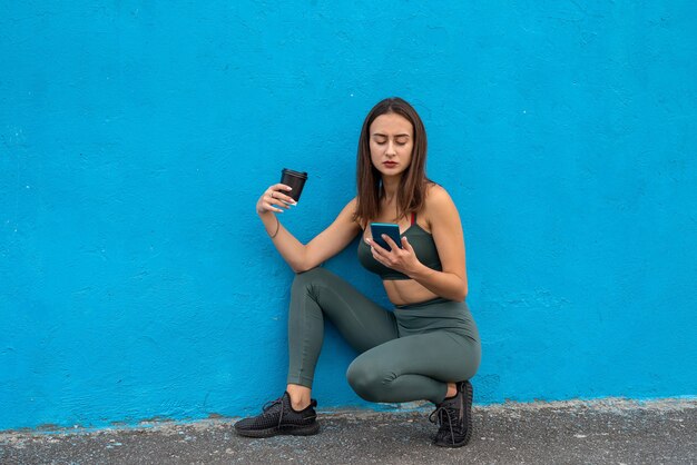 Mujer joven delgada en auriculares con una taza de bebida y escuchar música con el teléfono después de hacer ejercicio aislado sobre fondo azul.