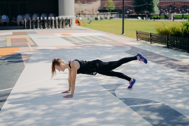 Mujer joven delgada deportiva hace ejercicio de tablaje levanta la pierna demuestra su determinación usa ropa deportiva y zapatillas de deporte entrena al aire libre escucha música en auriculares Concepto de estilo de vida saludable