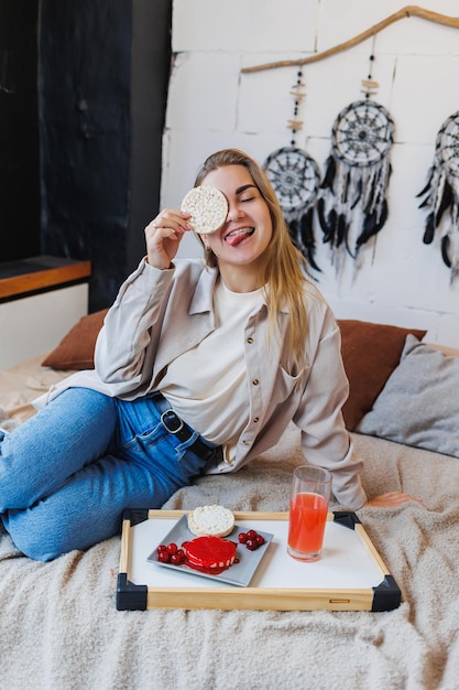 Una mujer joven y delgada está sentada en la cama y comiendo un delicioso desayuno de pasteles de arroz y jugo fresco Alimentación saludable