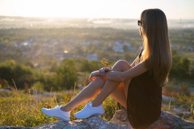 Mujer joven delgada en vestido corto negro sentado en una roca relajándose al aire libre al atardecer de verano. mujer de moda disfrutando de una cálida noche en la naturaleza.