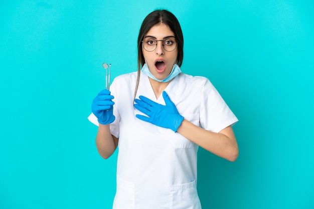 Mujer joven dentista caucásica sosteniendo herramientas aisladas sobre fondo azul sorprendido y conmocionado mientras mira a la derecha