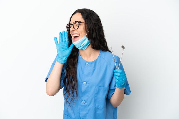 Mujer joven dentista sosteniendo herramientas aisladas sobre fondo blanco gritando con la boca abierta hacia el lado