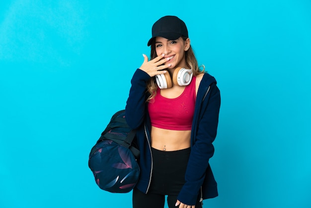 Mujer joven deporte con bolsa de deporte aislada en el espacio azul feliz y sonriente cubriendo la boca con la mano