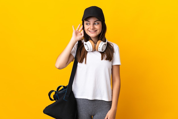 Mujer joven deporte con bolsa de deporte aislado en amarillo escuchando algo poniendo la mano en la oreja