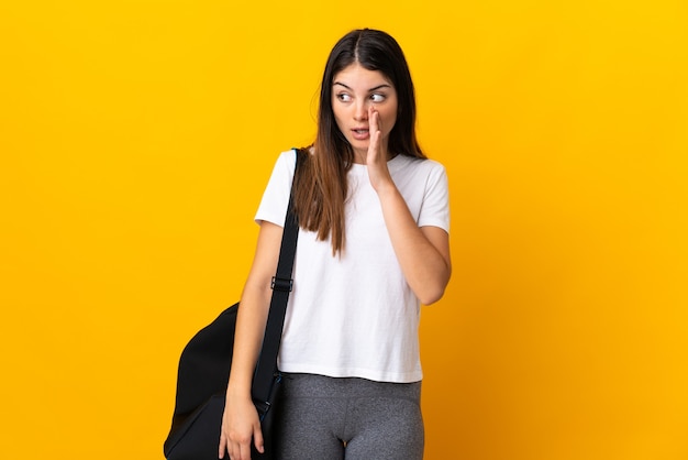 Mujer joven deporte con bolsa de deporte aislado en amarillo susurrando algo con gesto de sorpresa mientras mira hacia un lado