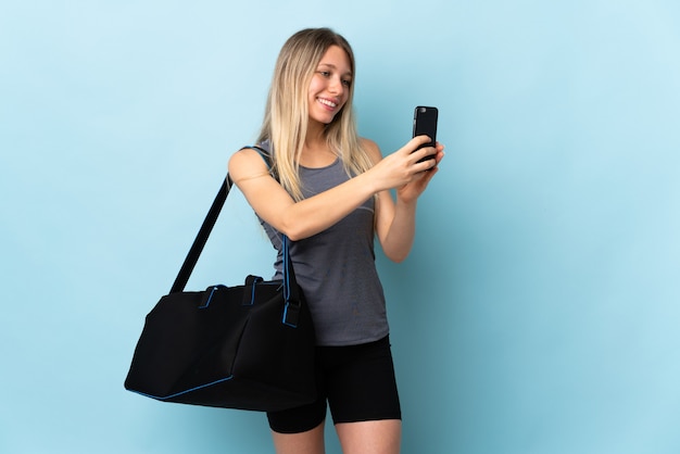 Mujer joven deporte con bolsa de deporte aislado en azul haciendo un selfie