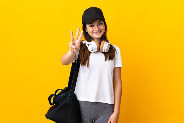 Mujer joven deporte con bolsa de deporte aislado en la pared amarilla feliz y contando tres con los dedos