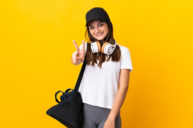 Mujer joven deporte con bolsa de deporte aislado en la pared amarilla sonriendo y mostrando el signo de la victoria