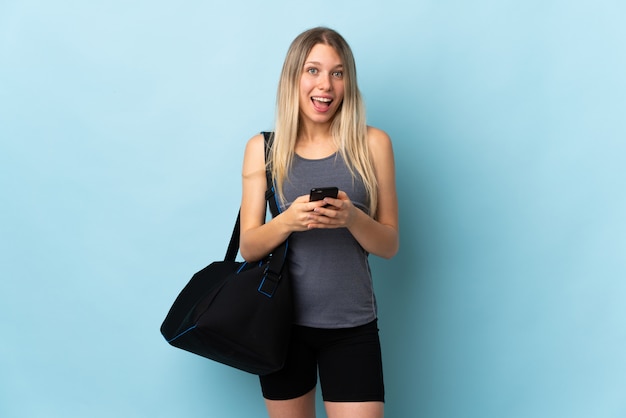 Mujer joven deporte con bolsa de deporte aislado en la pared azul sorprendido y enviando un mensaje