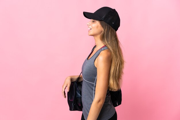 Mujer joven deporte con bolsa de deporte aislado sobre fondo rosa riendo en posición lateral